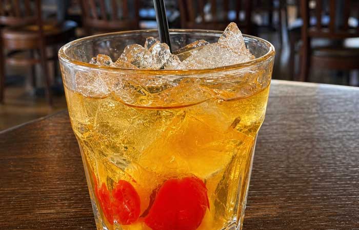 Glass of iced cocktail with two red cherries and a black straw from the bar menu Monticello MN.